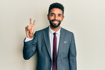 Handsome hispanic man with beard wearing business suit and tie smiling looking to the camera showing fingers doing victory sign. number two.