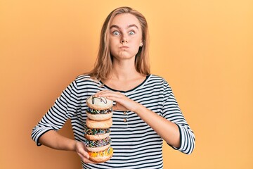 Beautiful blonde woman holding tasty colorful doughnuts puffing cheeks with funny face. mouth...