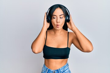 Naklejka premium Young beautiful hispanic girl listening to music using headphones making fish face with mouth and squinting eyes, crazy and comical.