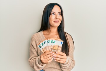 Young beautiful hispanic girl holding bunch of 50 euro banknotes smiling looking to the side and staring away thinking.