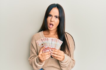 Young beautiful hispanic girl holding 10 colombian pesos banknotes in shock face, looking skeptical and sarcastic, surprised with open mouth