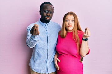 Young interracial couple expecting a baby, touching pregnant belly doing italian gesture with hand and fingers confident expression