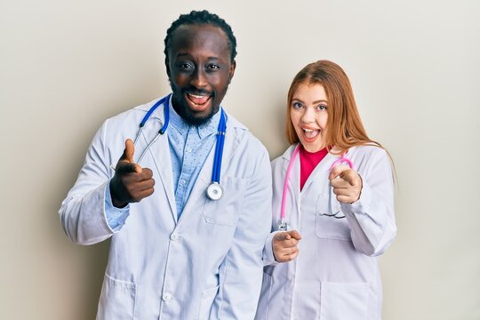 Young interracial couple wearing doctor uniform and stethoscope pointing fingers to camera with happy and funny face. good energy and vibes.
