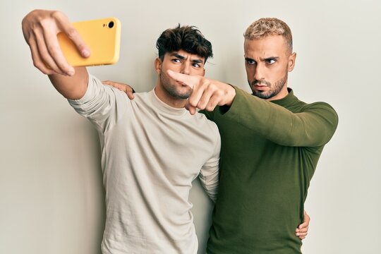 Homosexual gay couple standing together taking a selfie photo with smartphone pointing with finger to the camera and to you, confident gesture looking serious