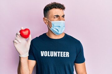 Hispanic young man wearing medical mask wearing volunteer t shirt holding heart angry and mad...