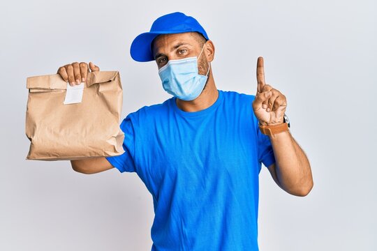 Handsome man with beard delivering food wearing covid-19 safety mask smiling with an idea or question pointing finger with happy face, number one
