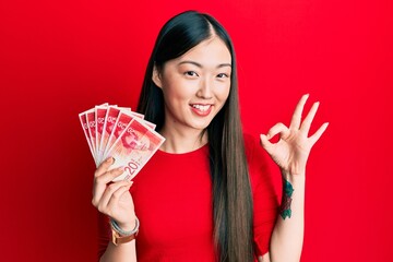 Young chinese woman holding 20 israel shekels banknotes doing ok sign with fingers, smiling friendly gesturing excellent symbol