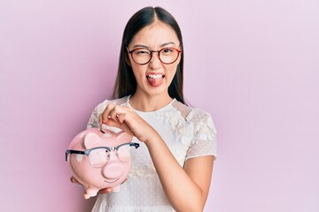 Young chinese woman holding piggy bank with glasses sticking tongue out happy with funny expression.