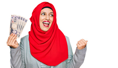 Beautiful hispanic woman wearing islamic hijab holding japanese yen banknotes pointing thumb up to the side smiling happy with open mouth