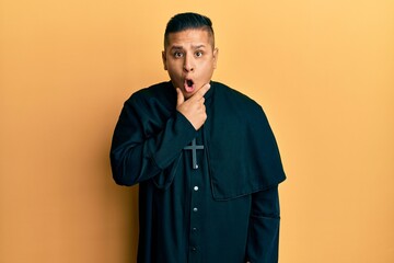 Young latin priest man standing over yellow background looking fascinated with disbelief, surprise and amazed expression with hands on chin