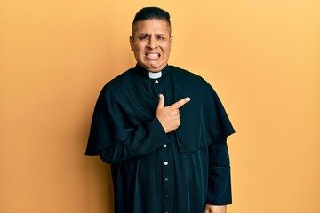 Young latin priest man standing over yellow background pointing aside worried and nervous with...
