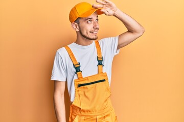 Hispanic young man wearing handyman uniform very happy and smiling looking far away with hand over head. searching concept.