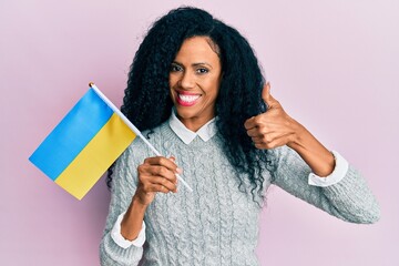 Middle age african american woman holding ukraine flag smiling happy and positive, thumb up doing excellent and approval sign