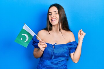 Young brunette teenager holding pakistan flag screaming proud, celebrating victory and success very excited with raised arm