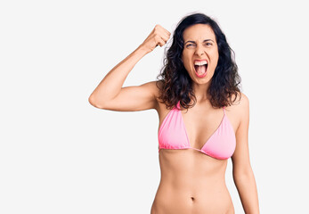 Young beautiful hispanic woman wearing bikini angry and mad raising fist frustrated and furious while shouting with anger. rage and aggressive concept.