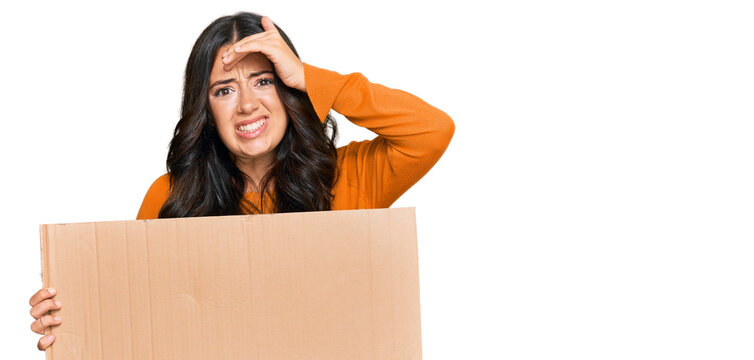 Beautiful brunette young woman holding cardboard blank empty banner stressed and frustrated with hand on head, surprised and angry face