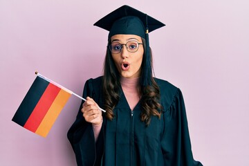Young hispanic woman wearing graduation uniform holding germany flag scared and amazed with open mouth for surprise, disbelief face