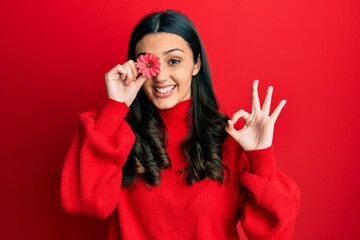 Young hispanic woman holding flower over eye doing ok sign with fingers, smiling friendly gesturing excellent symbol