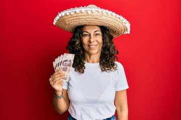 Middle age hispanic woman wearing mexican hat holding pesos banknotes looking positive and happy standing and smiling with a confident smile showing teeth