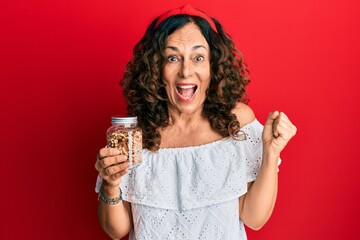 Middle age hispanic woman holding jar with walnuts screaming proud, celebrating victory and success very excited with raised arm