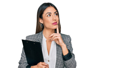 Young hispanic woman wearing business clothes holding clipboard serious face thinking about question with hand on chin, thoughtful about confusing idea