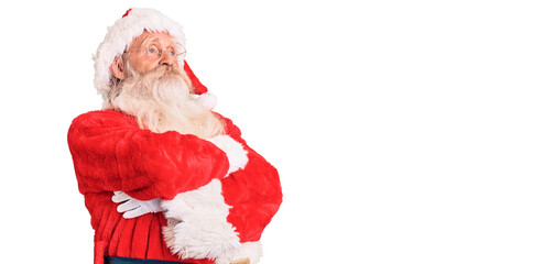 Old senior man with grey hair and long beard wearing traditional santa claus costume looking to the side with arms crossed convinced and confident
