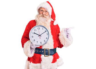 Old senior man with grey hair and long beard wearing santa claus costume holding clock smiling happy pointing with hand and finger to the side