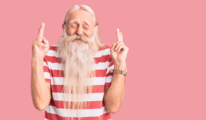Old senior man with grey hair and long beard wearing striped tshirt gesturing finger crossed smiling with hope and eyes closed. luck and superstitious concept.