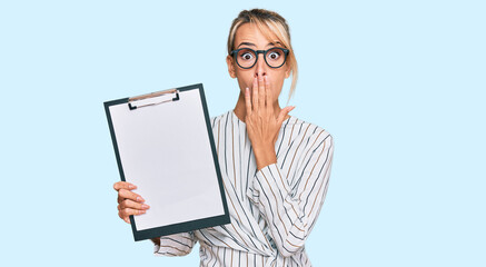 Beautiful blonde business woman holding clipboard with blank space covering mouth with hand,...
