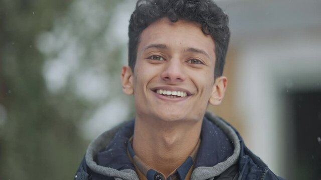 Close-up portrait of happy Middle Eastern male tourist standing outdoors on snowy day looking at camera smiling and breathing out. Headshot of confident positive young man posing on north.
