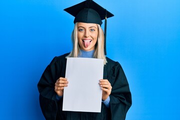 Beautiful blonde woman wearing graduation cap and ceremony robe holding blank banner sticking tongue out happy with funny expression.