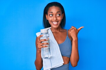 Young african american woman wearing sportswear drinking bottle of water pointing thumb up to the side smiling happy with open mouth