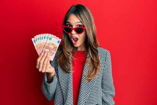 Young brunette woman holding bunch of 10 euro banknotes scared and amazed with open mouth for surprise, disbelief face