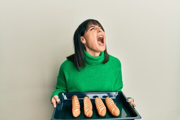 Young hispanic woman holding homemade bread angry and mad screaming frustrated and furious,...