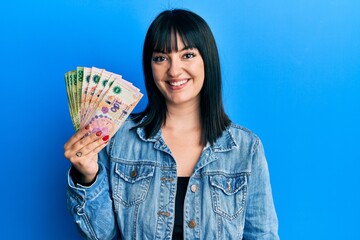 Young hispanic woman holding argentine pesos banknotes looking positive and happy standing and...