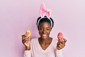 Young african american girl wearing cute easter bunny ears holding painted eggs smiling with a...