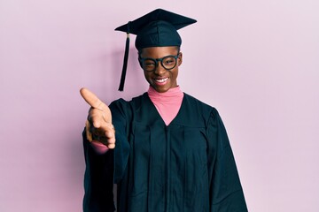 Young african american girl wearing graduation cap and ceremony robe smiling friendly offering handshake as greeting and welcoming. successful business.