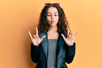 Teenager hispanic girl doing rock gesture making fish face with mouth and squinting eyes, crazy and comical.