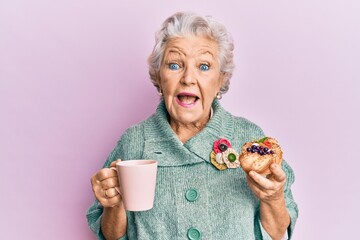 Senior grey-haired woman drinking a cup of coffee and eating bun celebrating crazy and amazed for...