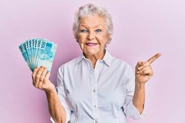 Senior grey-haired woman holding 100 brazilian real banknotes smiling happy pointing with hand and...