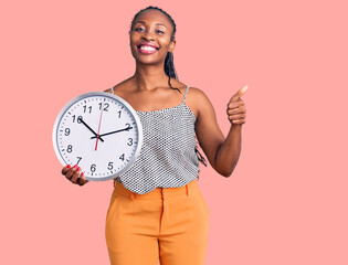 Young african american woman holding big clock smiling happy and positive, thumb up doing excellent and approval sign
