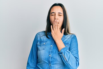 Young hispanic woman wearing casual denim jacket bored yawning tired covering mouth with hand....
