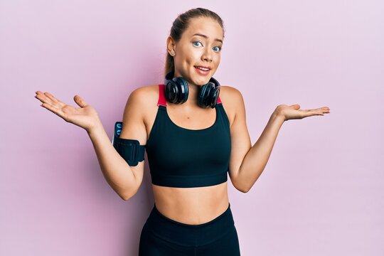 Beautiful young blonde woman wearing gym clothes and using headphones clueless and confused with open arms, no idea and doubtful face.