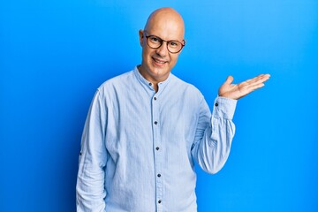 Middle age bald man wearing casual clothes and glasses smiling cheerful presenting and pointing with palm of hand looking at the camera.