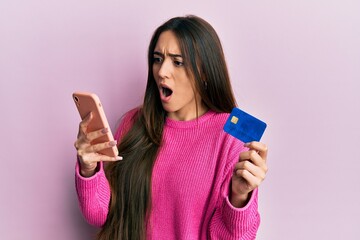 Young hispanic girl holding smartphone and credit card afraid and shocked with surprise and amazed expression, fear and excited face.