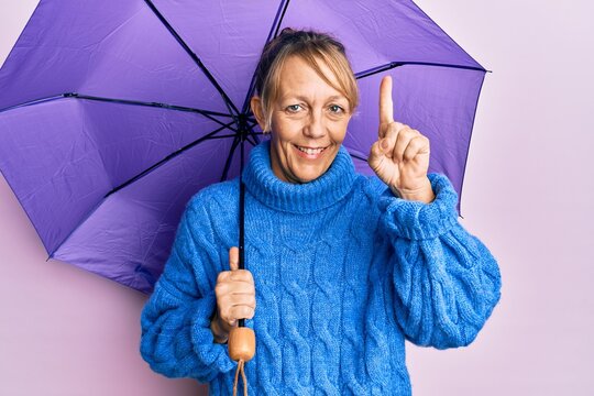 Middle age blonde woman holding purple umbrella smiling with an idea or question pointing finger with happy face, number one