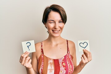 Young brunette woman with short hair holding heart and question mark reminder winking looking at...
