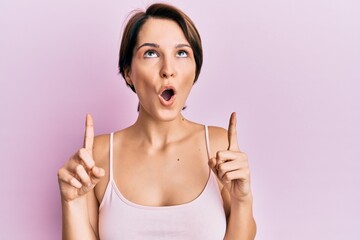 Young brunette woman with short hair over pink background amazed and surprised looking up and pointing with fingers and raised arms.