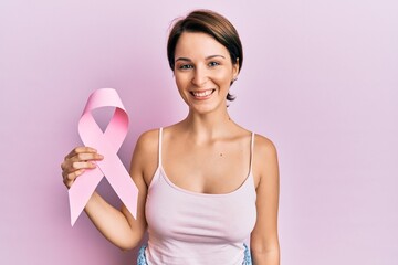 Obraz na płótnie Canvas Young brunette woman with short hair holding pink cancer ribbon looking positive and happy standing and smiling with a confident smile showing teeth