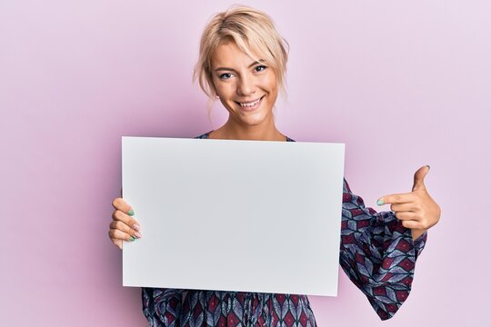 Young blonde girl holding blank empty banner smiling happy pointing with hand and finger